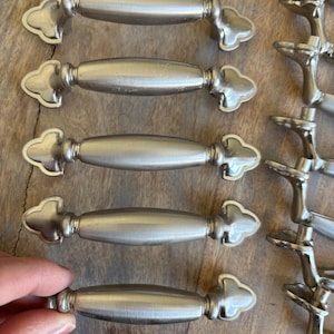 May include: Five silver cabinet knobs with a decorative, ornate design. The knobs have a slightly oval shape with a raised, rounded edge. The knobs are arranged in a row, with the top knob slightly offset from the others.