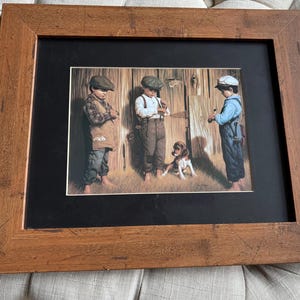 May include: A framed print of a painting depicting three boys standing in front of a wooden barn door. The boys are wearing caps and are holding various items. A small dog is standing in front of the boys. The print is framed in a brown wooden frame.