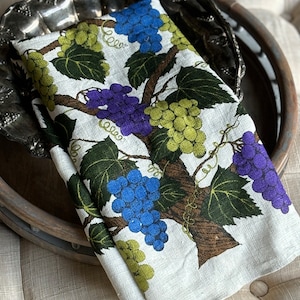May include: A white linen tea towel with a grapevine design. The grapes are green, purple, and blue. The leaves are green and the vine is brown.