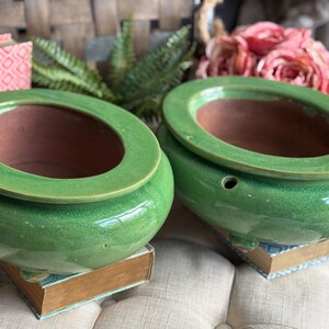 May include: Two green ceramic planters with a glossy finish. Each planter has a wide, circular rim and a brown interior. They sit atop vintage books, with a blurred background of flowers and greenery.