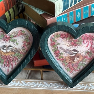 May include: Two heart-shaped wall decor pieces with a dark green frame. Each features a bird illustration within a floral wreath. The background includes books and greenery.