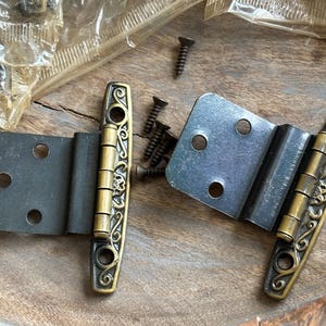 May include: Two ornate brass hinges with a black metal base. The hinges have a decorative scroll design and are attached to a wooden surface.