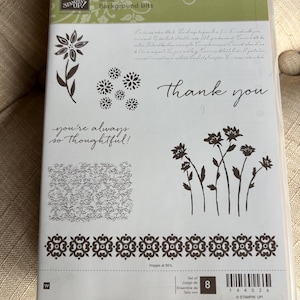 May include: Clear plastic case containing a set of rubber stamps. The stamps feature floral designs, the words "thank you", and "you're always so thoughtful!" in a handwritten font. The set is labeled "Background Bits".