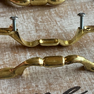 May include: Two gold-toned metal drawer pulls with a curved, elongated shape and a central rectangular section. One pull is shown from the side with two silver-colored mounting screws attached, while the other pull is positioned below it.