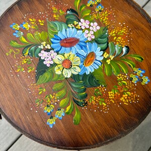 May include: A round wooden table top with a hand-painted floral design. The design features blue, yellow, white, and green flowers with orange centres. The flowers are surrounded by green leaves and small yellow and orange dots.