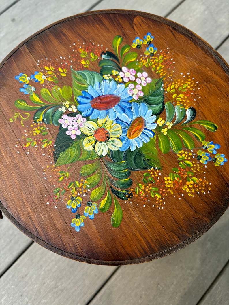May include: A round wooden table top with a hand-painted floral design. The design features blue, yellow, white, and green flowers with orange centres. The flowers are surrounded by green leaves and small yellow and orange dots.
