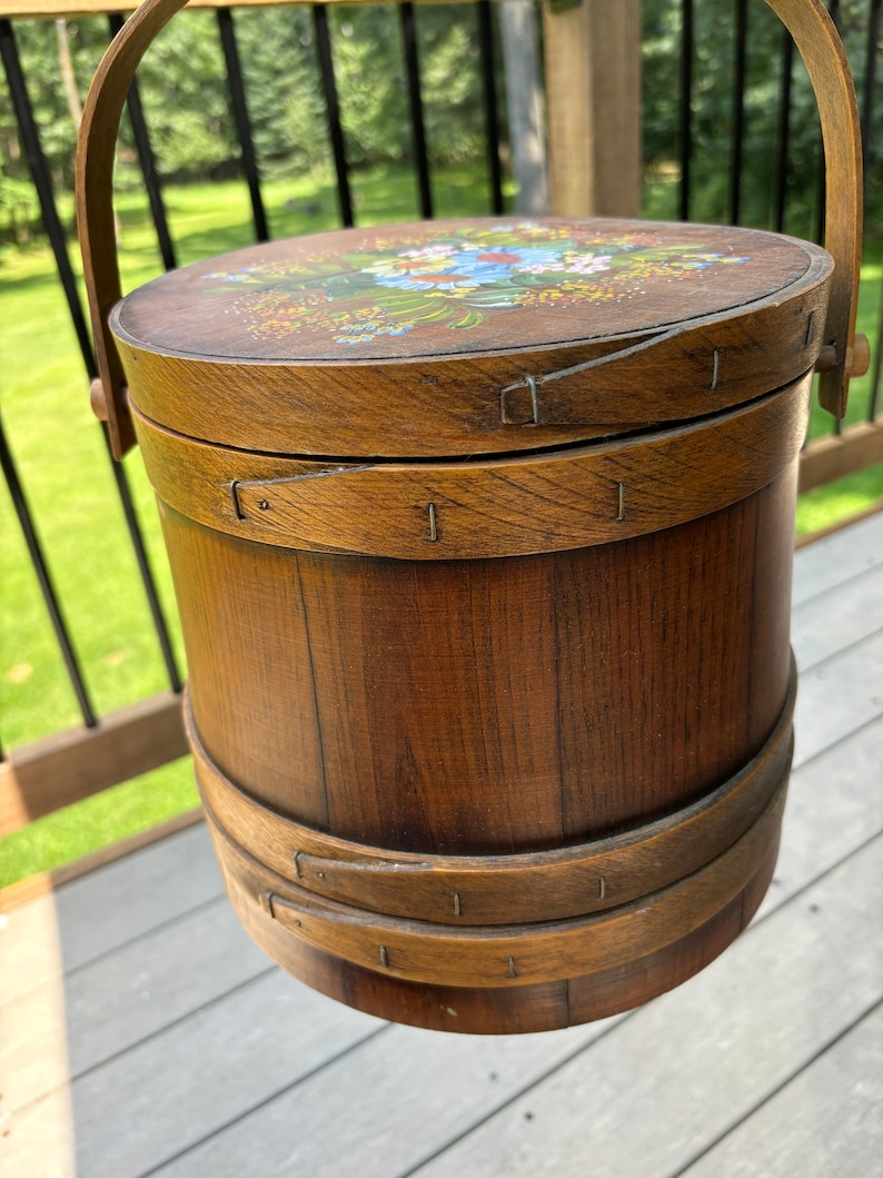 May include: A wooden bucket with a painted floral design on the lid. The bucket has a handle and is made of natural wood.