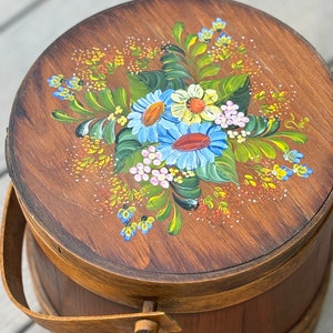 May include: A wooden bucket with a painted floral design on the lid. The flowers are blue, yellow, pink, and green. The bucket has a wooden handle.