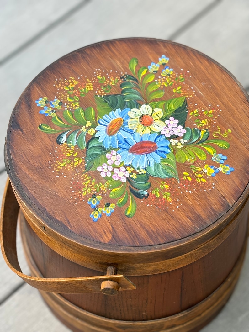 May include: A wooden bucket with a painted floral design on the lid. The flowers are blue, yellow, pink, and green. The bucket has a wooden handle.