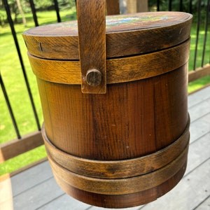 May include: A wooden bucket with a handle and a painted floral design on the lid. The bucket is brown with a dark brown stain and has a metal handle.