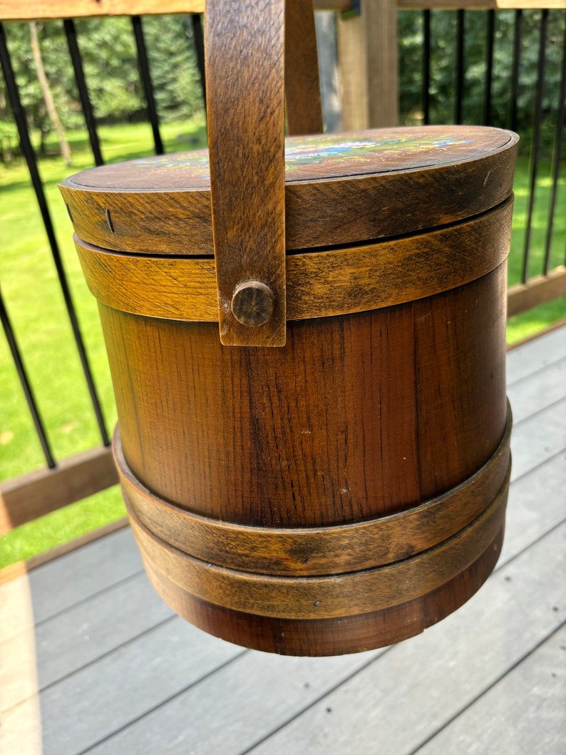 May include: A wooden bucket with a handle and a painted floral design on the lid. The bucket is brown with a dark brown stain and has a metal handle.