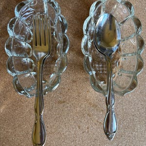 May include: Two clear glass spoon rests with a textured, scalloped design. Each rest holds a piece of silverware: a fork and a spoon. The silverware has a decorative handle design. The items are displayed on a neutral surface.