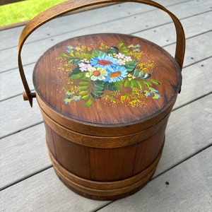 May include: A wooden bucket with a handle, painted with a floral design in shades of blue, yellow, green, and pink. The bucket is made of wood and has a natural brown finish.