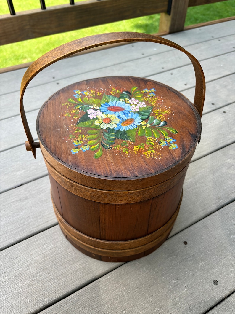 May include: A wooden bucket with a handle, painted with a floral design in shades of blue, yellow, green, and pink. The bucket is made of wood and has a natural brown finish.