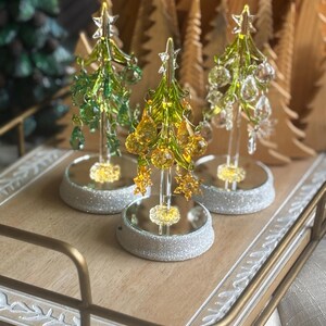 May include: Three decorative glass Christmas trees with crystal-like ornaments and star toppers sit on a mirrored base with a glittery silver border. The trees are displayed on a wooden tray with gold handles.