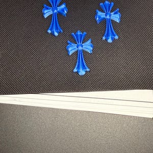 May include: Three blue cross-shaped ornaments with a textured appearance are arranged on a dark surface. Each cross has a detailed design with a central point and four outward-facing arms, creating a decorative element.