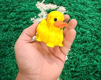 May include: A yellow rubber duck with an orange beak and black eyes. The duck is held in a hand against a green and white background.