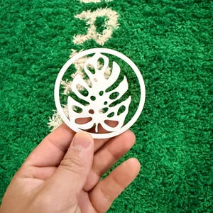 Puede incluir: Una pieza decorativa blanca y circular con un diseño de hoja de monstera estilizada. La pieza se sostiene sobre un fondo verde texturizado con texto blanco. El diseño es un recorte.