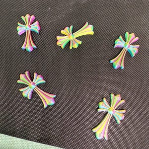 May include: Five colorful cross-shaped embellishments in shades of pink, yellow, green, and blue. The crosses have a textured appearance and are arranged on a dark gray surface. These decorative elements could be used for crafts or jewelry making.