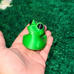 May include: A bright green frog-shaped figurine with black eyes. The frog is a solid color and has a smooth texture. The figurine is held in a hand against a green background. This item is a decorative piece.