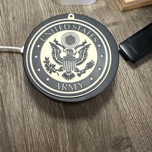 以下が含まれることがあります： アメリカ陸軍のエンブレムが付いた白黒のワイヤレス充電器。充電器は円形で、「United States Army」の文字がエンブレムを囲んでいます。充電器の直径は約10cmです。
