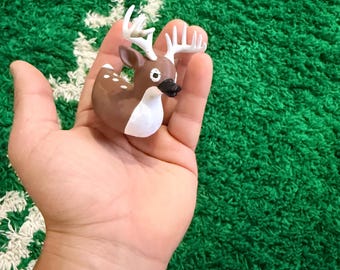 Puede incluir: Una pequeña figura de ciervo marrón y blanco con cuernos blancos y una nariz negra. El ciervo tiene manchas blancas en la espalda y se sostiene en una mano. El fondo es una superficie verde texturizada.