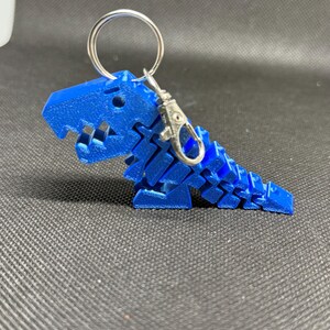 May include: A blue, 3D-printed dinosaur keychain. The dinosaur is segmented, with a detailed head and body. It has a silver keyring and clasp for attaching to keys or a bag. The keychain is made of plastic.
