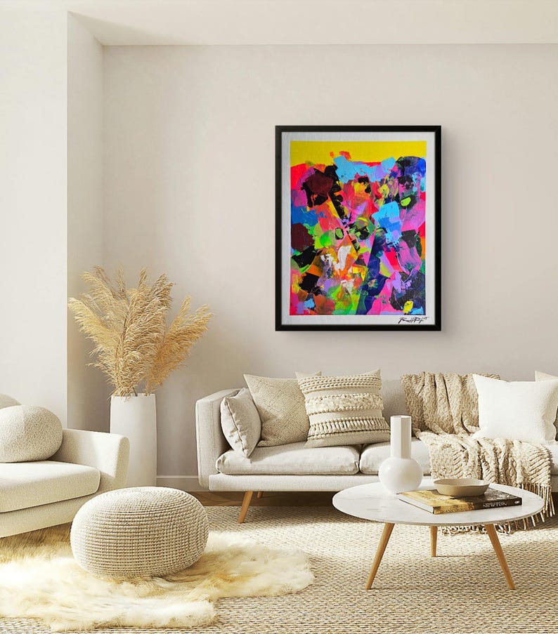 May include: Abstract art print in a black frame. The artwork features a vibrant mix of colours, including yellow, pink, blue, and green, with bold brushstrokes. The painting is signed in the bottom right corner. The artwork is displayed in a living room setting.