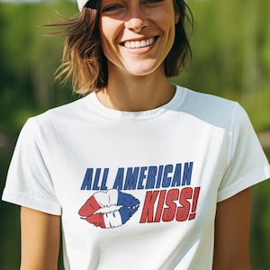 May include: White t-shirt with the text "ALL AMERICAN KISS!" in blue and red, along with a graphic of lips in red, white, and blue. The person is wearing a white cap and smiling.