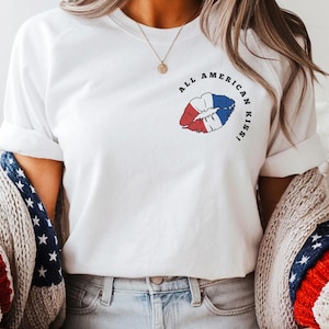 May include: White t-shirt with a graphic of lips in red, white, and blue, with the text "ALL AMERICAN KISS!". The person is wearing denim shorts, brown cowboy boots, and a sweater with red, white, and blue stripes and stars.