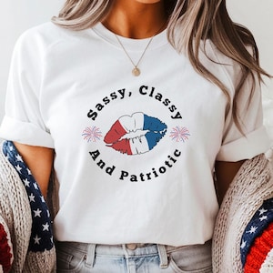 May include: White t-shirt with the text "Sassy, Classy, And Patriotic" surrounding a graphic of lips in red, white, and blue. The shirt is paired with denim shorts and brown cowboy boots. A patriotic-themed sweater is draped over the shoulders.