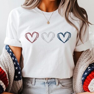 May include: White t-shirt featuring three outlined hearts in red, white, and blue. The shirt is paired with denim shorts, a patriotic sweater, and brown cowboy boots. The overall aesthetic is casual and Americana-themed.