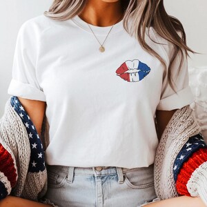 May include: White t-shirt with a red, white, and blue lip print. The person is wearing denim shorts, brown cowboy boots, and a gray sweater with red, white, and blue stripes and stars.