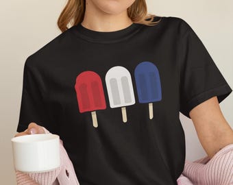 May include: Black t-shirt featuring a graphic of three popsicle treats in red, white, and blue. The popsicles are on wooden sticks. The shirt is a crew neck and has short sleeves.