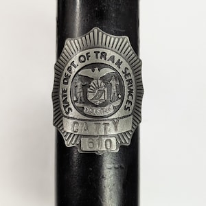 Puede incluir: Objeto de metal negro con un emblema plateado. El emblema dice "STATE DEPT. OF TRAM SERVICES DELSINO CATTY 610" en un diseño circular con un águila y una escena de montaña.
