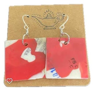 May include: Square red and white earrings with silver hooks. The earrings are attached to a brown card with a drawing of a genie lamp. The earrings have a unique, abstract design.