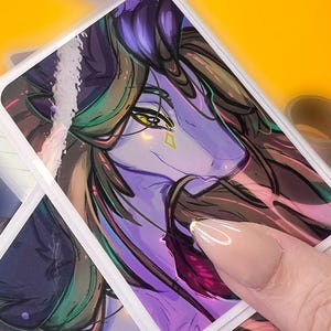 May include: A stack of rectangular stickers featuring a purple and green fantasy creature with yellow eyes and a diamond-shaped marking. The stickers have a white border and are held by a hand with a light-colored nail. The background is yellow.