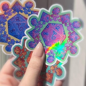 May include: Two colorful, holographic stickers featuring a central twenty-sided die surrounded by smaller dice. The stickers have a floral-like design with a variety of colors, including blue, purple, orange, and green. The stickers are designed for role-playing games.