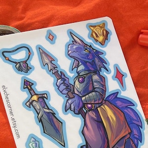 May include: A sticker sheet featuring a blue dragon-like character in armor holding a spear, accompanied by a sword, a potion bottle, and a necklace. The sheet includes the text elvchescorner.etsy.com and printed in canada, alongside a blank white backing sheet.