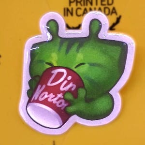 May include: A green cartoon character sticker with a white outline, holding a red and white can that says "Din Nortor." The sticker is on a yellow background with text and symbols.