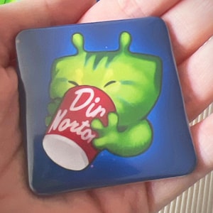 Könnte beinhalten: Quadratischer Magnet mit einem grünen Alien-Cartoon-Charakter, der eine rot-weiße "Dir Norton"-Tasse auf blauem Hintergrund hält. Weitere Magnete mit ähnlichen Charakteren sind sichtbar.