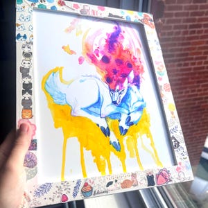 May include: Watercolor artwork of a white wolf with blue and purple accents, framed in a white frame decorated with cat stickers. The artwork features a yellow and orange background with a colorful, fiery mane.