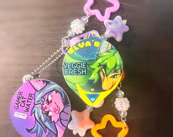 Op de afbeelding: Een decoratieve sleutelhanger met twee ronde bedels met anime-stijl illustraties. Eén bedel toont ELVA'S VEGGIE FRESH, de andere GAMER CAT WATER. De bedels zijn verbonden door een zilveren kogelketting met roze, oranje en paarse stervormige kralen.