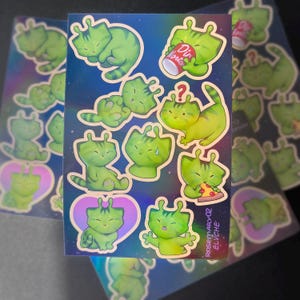 Op de afbeelding: Vel holografische stickers met groene alien kat illustraties in verschillende poses. Sommige stickers tonen de kat met een drankje, pizza of een vraagteken. De achtergrond heeft een ruimtethema.