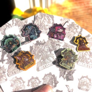 May include: A collection of six small, colorful enamel pins featuring stylized creature illustrations. Each pin displays a specific label including OCD, PTSD, Anxiety, Autism, and Depression, set against a white backing card with repeating patterns.