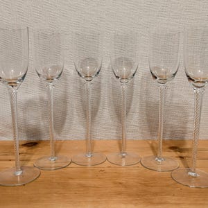 May include: Six clear champagne flutes with long, twisted stems and round bases are arranged on a wooden surface. The glasses have a classic, elegant design, perfect for celebratory occasions. The stems have a textured, rope-like appearance.