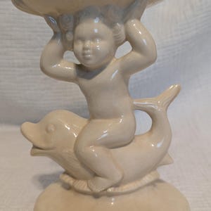 Puede incluir: Una figurita de cerámica blanca de un querubín montando un delfín, sosteniendo un plato de cerámica blanca sobre su cabeza.