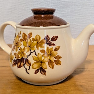 以下が含まれることがあります： 茶色の蓋と花柄の白い陶器製のティーポット。ティーポットには黄色と茶色の花と緑の葉が描かれています。
