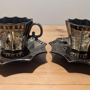 May include: Two black ceramic teacups and matching saucers featuring gold-coloured Egyptian-themed motifs, including hieroglyphics and pharaonic imagery. The cups have a scalloped rim and a curved handle, while the saucers have a matching octagonal shape.