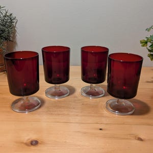 May include: Four ruby red stemmed glasses with clear bases are arranged on a wooden surface. The glasses have a cylindrical shape with a rounded base. The background includes plants in pots.